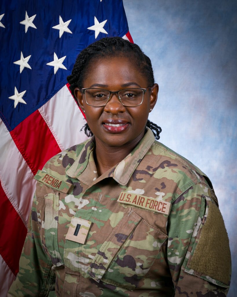 Portrait of a U.S. Air Force service member in camouflage uniform wearing glasses, standing in front of an American flag.